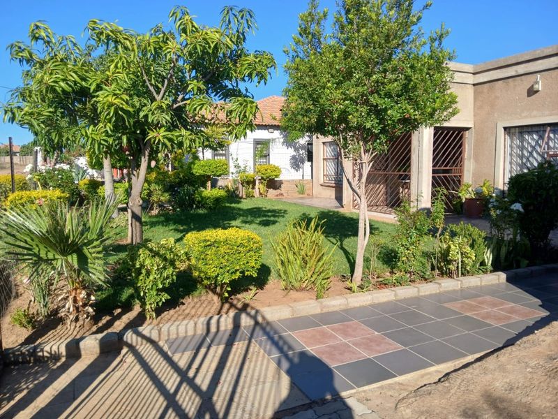 A well-maintained garden area featuring a manicured lawn, established trees, and a paved patio section with security gate access to the home.