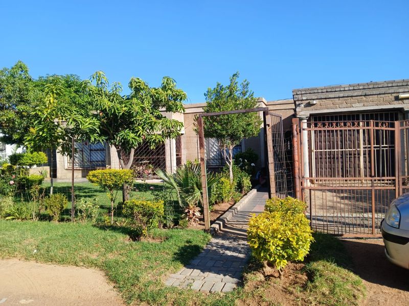 Front view of the property featuring a paved walkway, security gate, and established garden area with brick facade and security bars on windows.