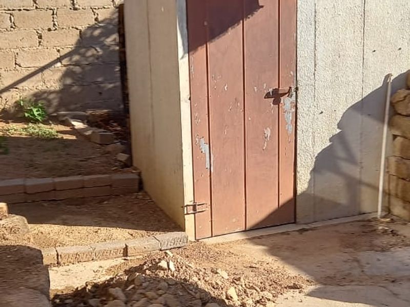 A small, detached utility structure with a metal door, situated in a yard area with concrete flooring and surrounding block walling.