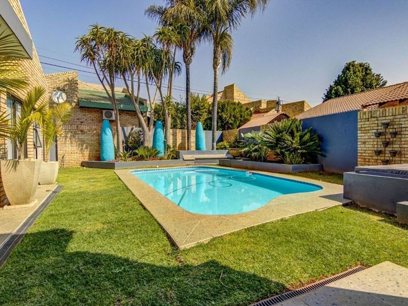 A well-maintained outdoor pool area featuring a clear swimming pool, paved surround, and established garden landscaping within a private brick-walled enclosure.