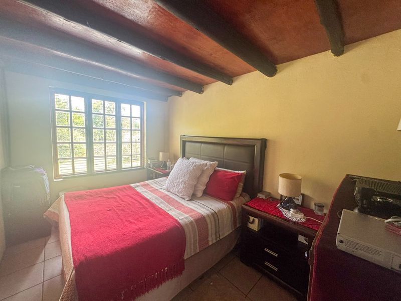 A cozy bedroom featuring exposed timber ceiling beams, tiled flooring, and a multi-pane window providing natural light.