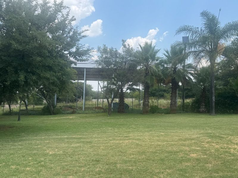 A spacious, well-maintained lawn area featuring mature trees and a large open-sided metal frame structure in the background.
