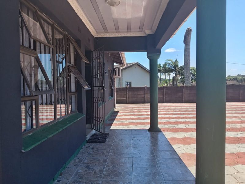 A covered patio area featuring tiled flooring and support columns, overlooking a paved driveway and perimeter wall.