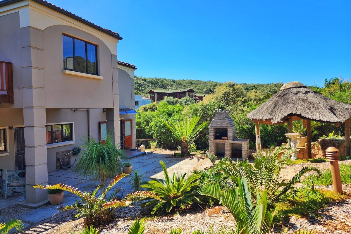 Rear exterior view featuring a paved patio area, a built-in brick braai, and a thatched lapa set within a landscaped garden.