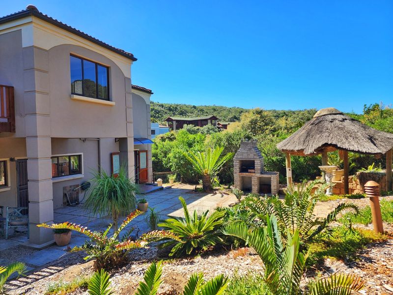 Rear exterior view featuring a paved patio area, a built-in brick braai, and a thatched lapa set within a landscaped garden.