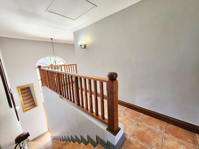A bright upper-level hallway featuring a wooden balustrade, ceramic tiled flooring, and an arched window overlooking the staircase area.