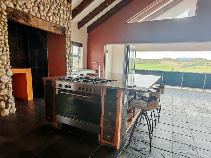 Spacious kitchen featuring a stone-clad fireplace, exposed timber beams, and a central island with a gas stove, opening onto a scenic outdoor patio.