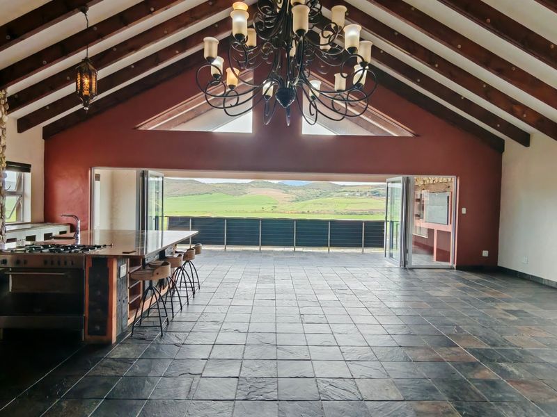 Spacious open-plan living and kitchen area featuring vaulted ceilings with exposed beams, slate flooring, and large bi-fold doors opening to scenic landscape views.