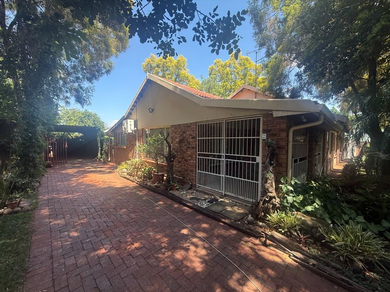 Front elevation of a brick house featuring a paved driveway, security gates, and established garden surroundings.