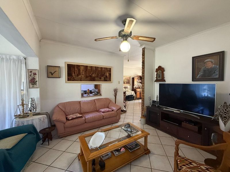 A comfortable living room featuring ceramic floor tiles, a ceiling fan, and a brick feature pillar, with a convenient serving hatch to the kitchen area.