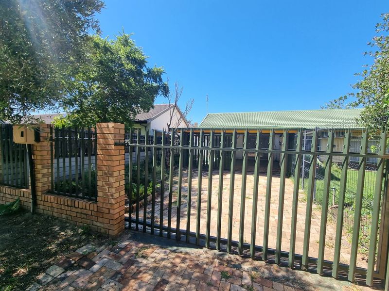 Front view of the property featuring a paved driveway, secure metal gate, and brick perimeter wall with established garden surroundings.