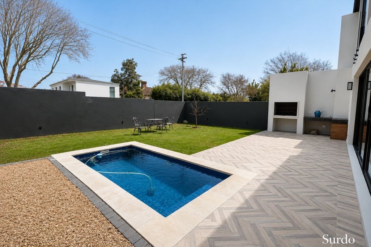 A well-maintained rear garden featuring a swimming pool, herringbone paved patio, and a built-in outdoor braai area against a modern boundary wall.