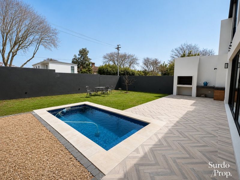A well-maintained rear garden featuring a swimming pool, herringbone paved patio, and a built-in outdoor braai area against a modern boundary wall.