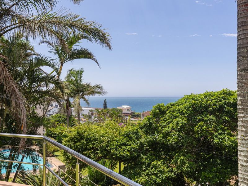 Elevated view overlooking the surrounding coastal neighborhood, lush greenery, and the ocean horizon, captured from a balcony with stainless steel railings.