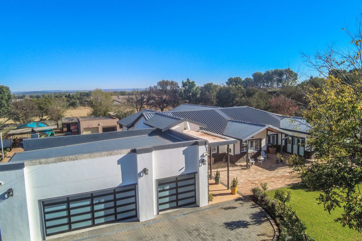 Aerial view of a modern single-story residence featuring a metal roof, double garage, expansive paved patio area, and well-maintained surrounding garden.