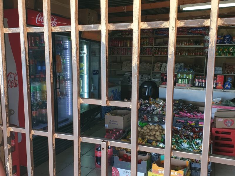 A small retail or utility space secured by a metal gate, featuring tiled floors, corrugated metal ceiling, and built-in shelving units.