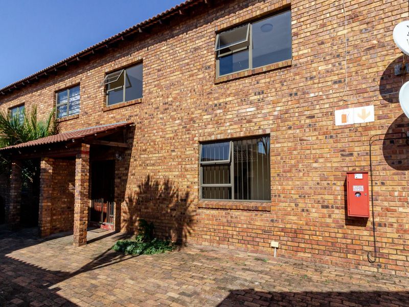A well-maintained face-brick townhouse exterior featuring a paved entrance area, covered porch, and secure aluminium-framed windows.