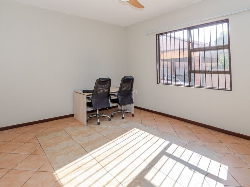A functional office space featuring ceramic floor tiles, a ceiling fan, and a window fitted with security bars.