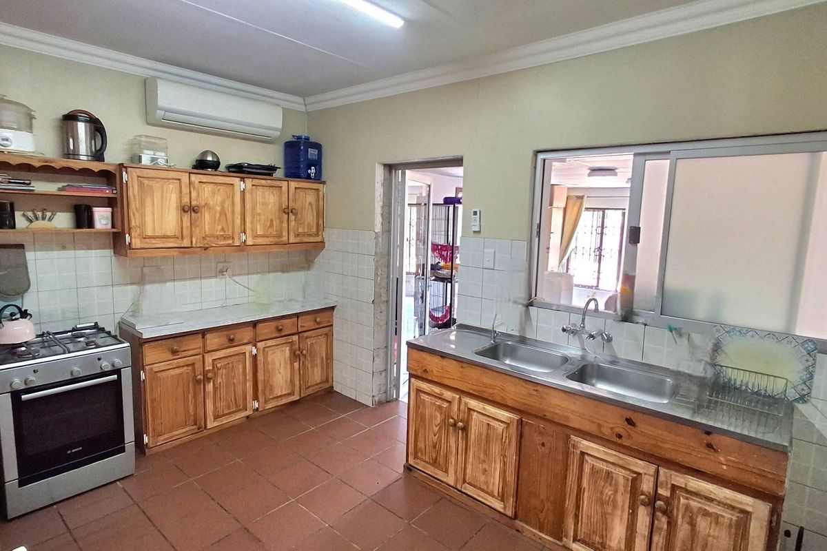 A functional kitchen featuring wooden cabinetry, a double stainless steel sink, tiled backsplash, and tiled flooring, with an integrated wall-mounted air conditioning unit.