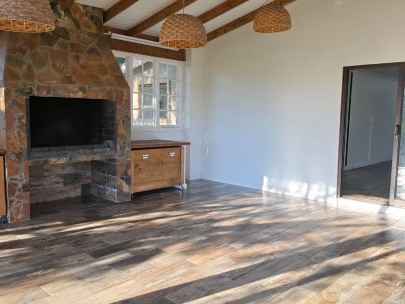 A spacious covered patio featuring a stone fireplace, exposed timber ceiling beams, and tiled flooring, offering a bright and versatile outdoor living area.