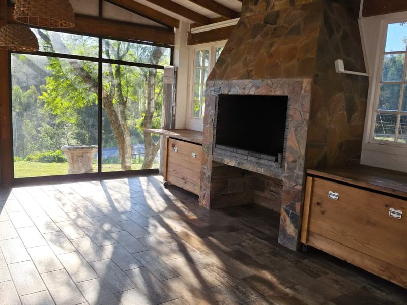 A bright, enclosed patio featuring a prominent stone-clad fireplace, built-in braai, wood-look tiled flooring, and expansive glass doors overlooking the garden.