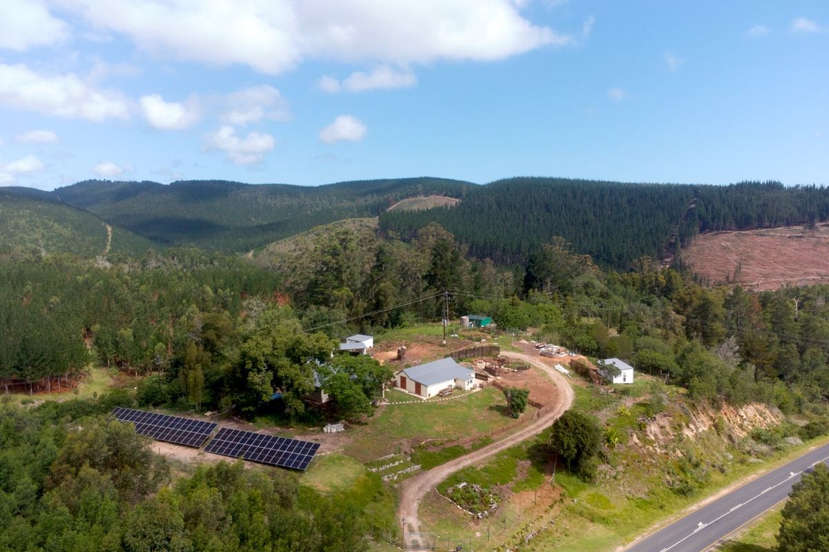 Aerial view of a rural farm property featuring multiple buildings, a large solar panel array, and a gravel driveway surrounded by forest landscape.