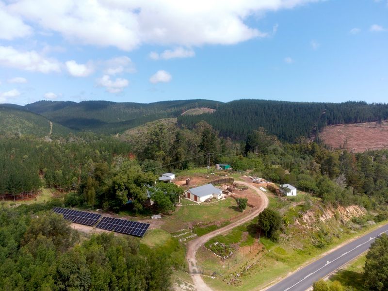 Aerial view of a rural farm property featuring multiple buildings, a large solar panel array, and a gravel driveway surrounded by forest landscape.