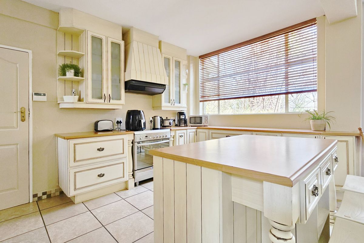 A traditional-style kitchen featuring cream-colored cabinetry, a central island with a wooden countertop, tiled flooring, and a large window with venetian blinds.