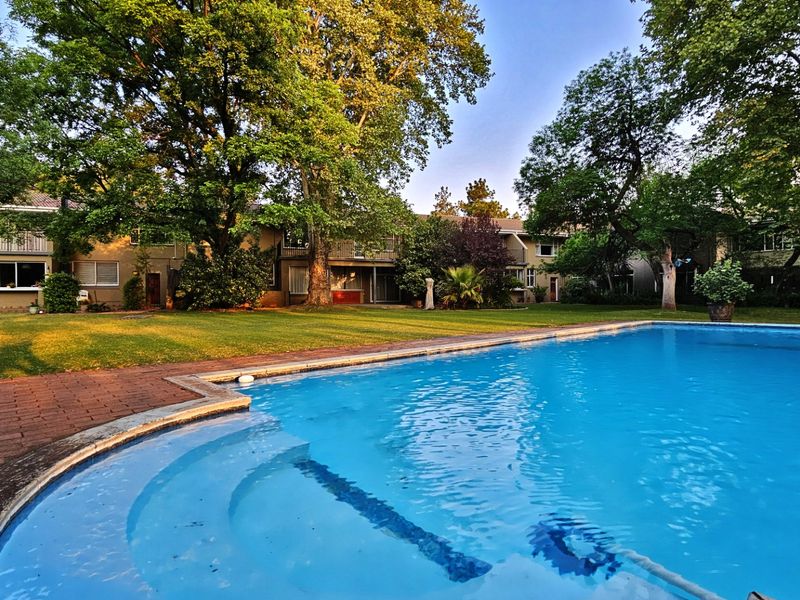 A communal swimming pool area set within a lush, landscaped garden featuring mature trees and brick paved surrounds, serving the townhouse complex.