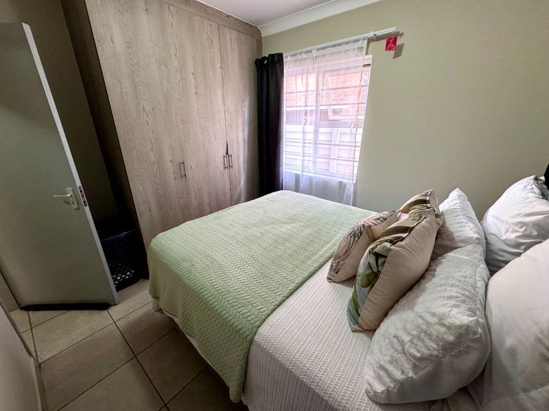 A functional bedroom featuring built-in wooden cupboards, tiled flooring, and a window fitted with security bars.