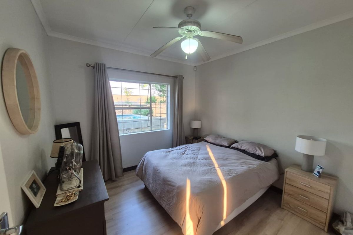 A well-maintained bedroom featuring laminate flooring, a ceiling fan, and a window with security bars overlooking the exterior pool area.