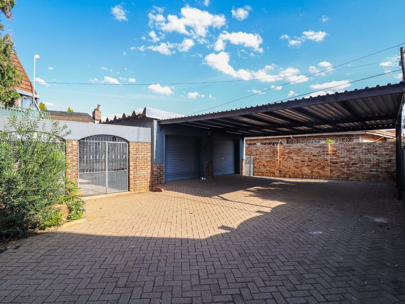A spacious paved driveway featuring a double carport and two secure roller shutter garage doors, enclosed by a brick boundary wall with security fencing.