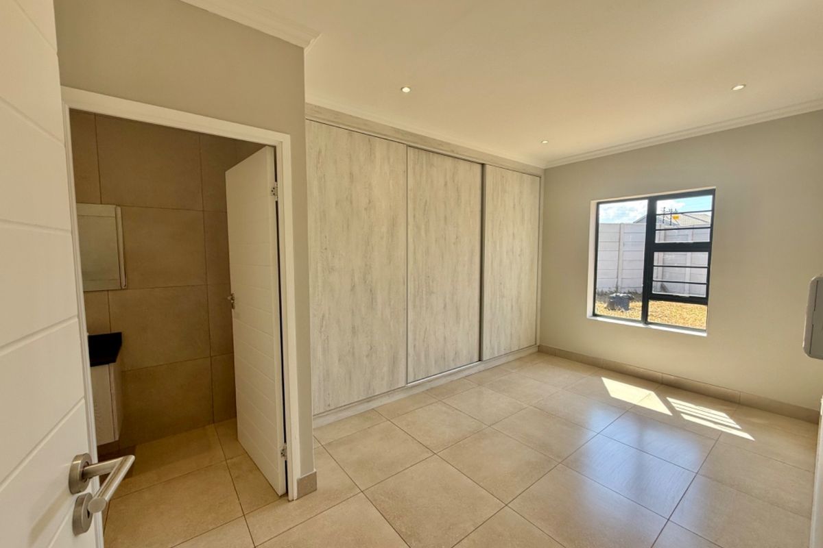 A bright, modern bedroom featuring neutral tiled flooring, built-in sliding door cupboards, recessed lighting, and an adjoining en-suite bathroom.