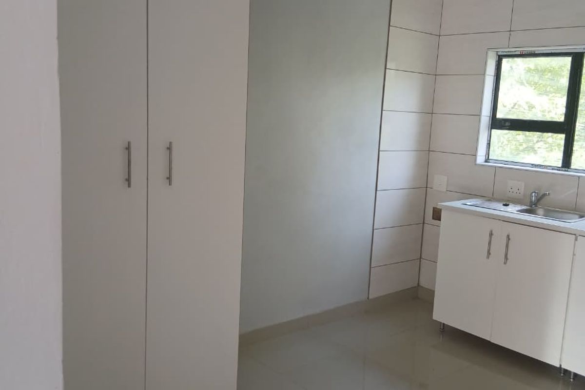 A clean, compact kitchen area featuring white built-in cabinetry, a stainless steel sink, tiled splashbacks, and neutral floor tiles.
