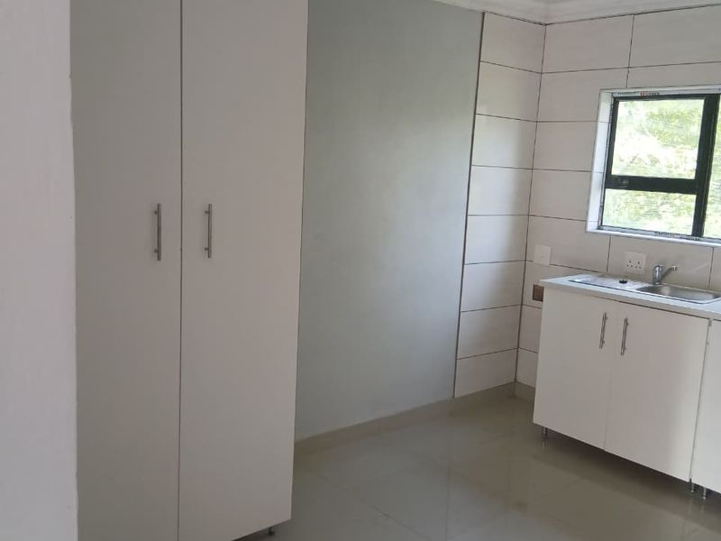 A clean, compact kitchen area featuring white built-in cabinetry, a stainless steel sink, tiled splashbacks, and neutral floor tiles.