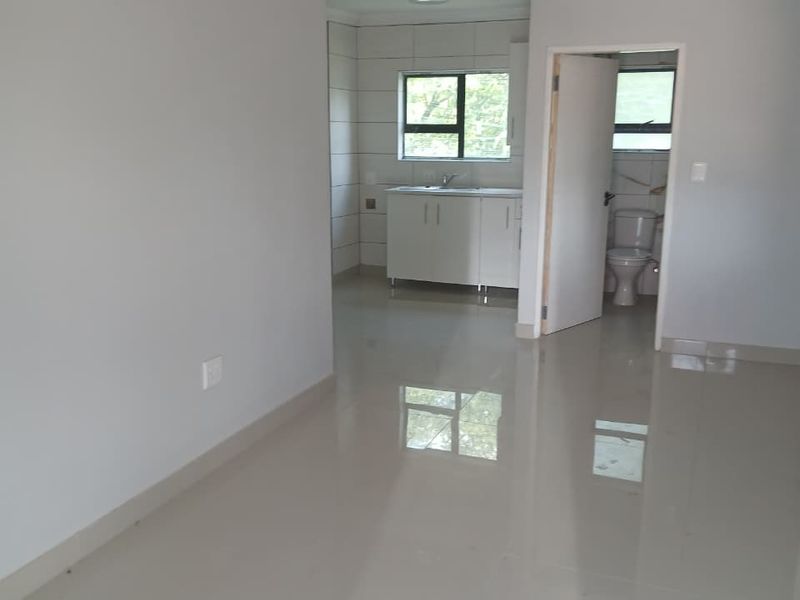 An open-plan living area featuring neutral tiled flooring, leading to a functional kitchen space and an adjacent bathroom with a toilet.