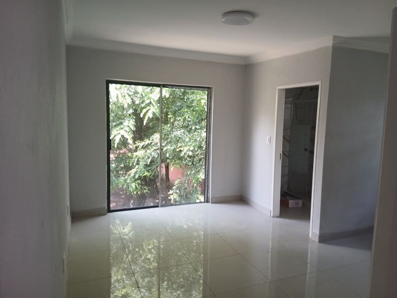 A bright, empty bedroom featuring neutral tiled flooring, a sliding glass door with garden views, and clean white walls.