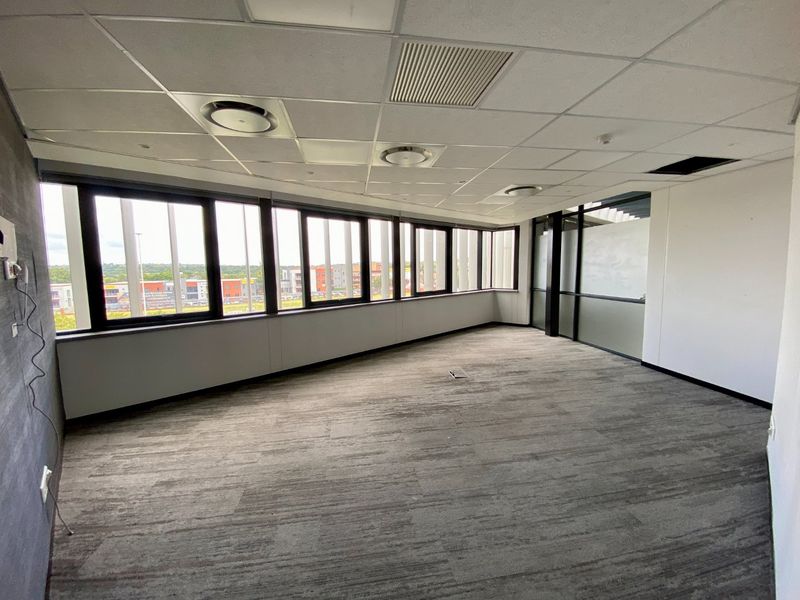 A bright, empty commercial office space featuring large windows, carpeted flooring, and a suspended ceiling with integrated lighting and air conditioning.