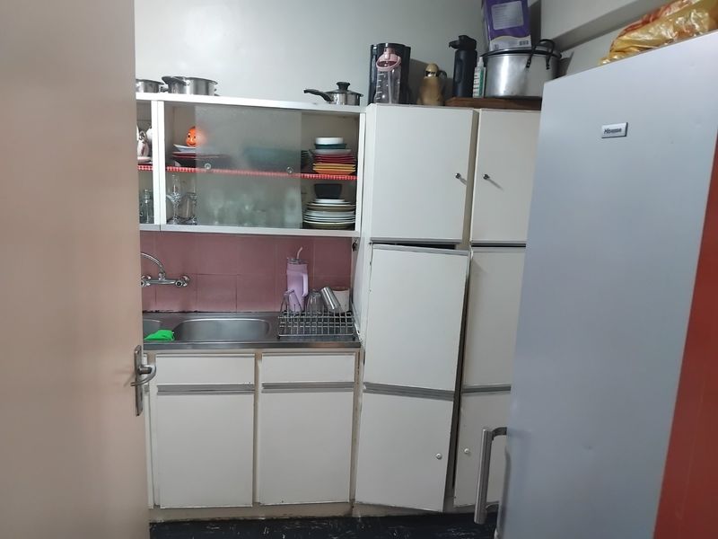 A compact kitchen featuring white built-in cabinetry, a stainless steel sink, and a pink tiled backsplash, showing signs of age and wear.