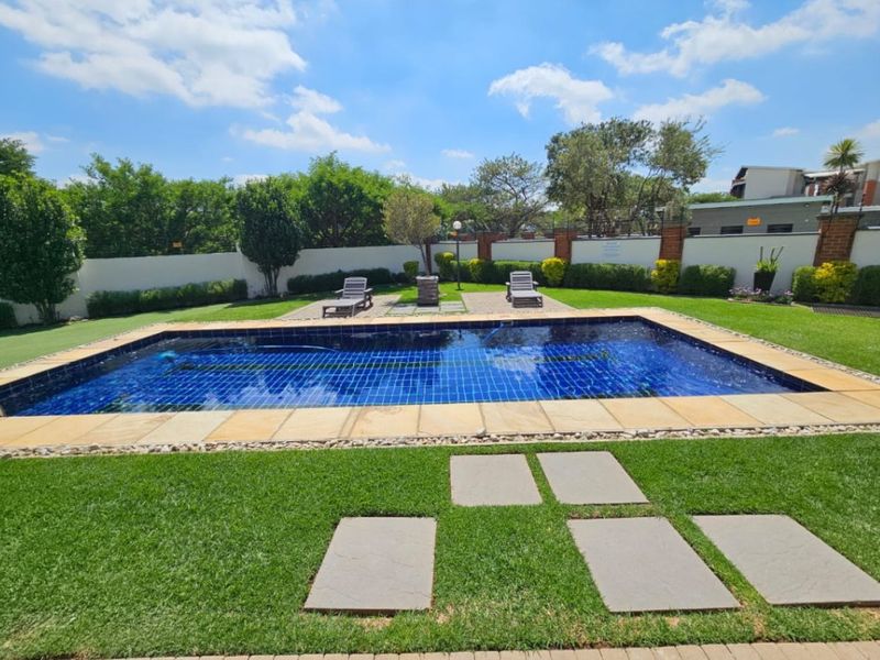 A well-maintained outdoor swimming pool area featuring stone coping, surrounding lawn, and a paved patio section enclosed by a perimeter wall.