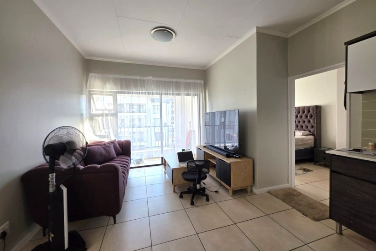 A bright living room featuring tiled flooring and a sliding glass door leading to a balcony, with an adjacent view into the bedroom.