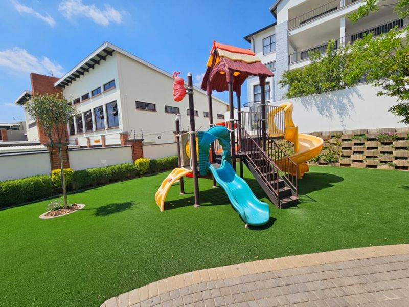 A well-maintained communal garden area featuring artificial turf, a children's playground structure, and secure perimeter walling with electric fencing.