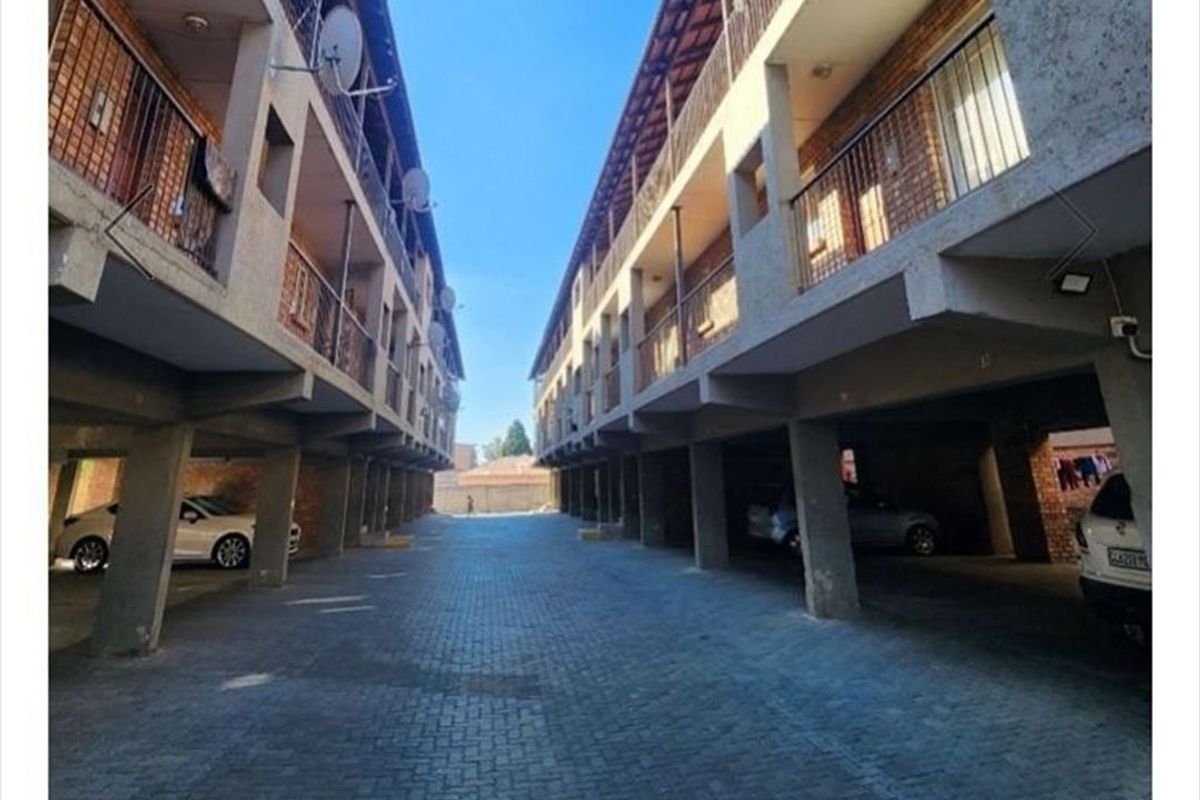 Exterior view of an apartment complex featuring a paved driveway, covered parking area beneath the building, and multiple levels of balconies with metal railings.