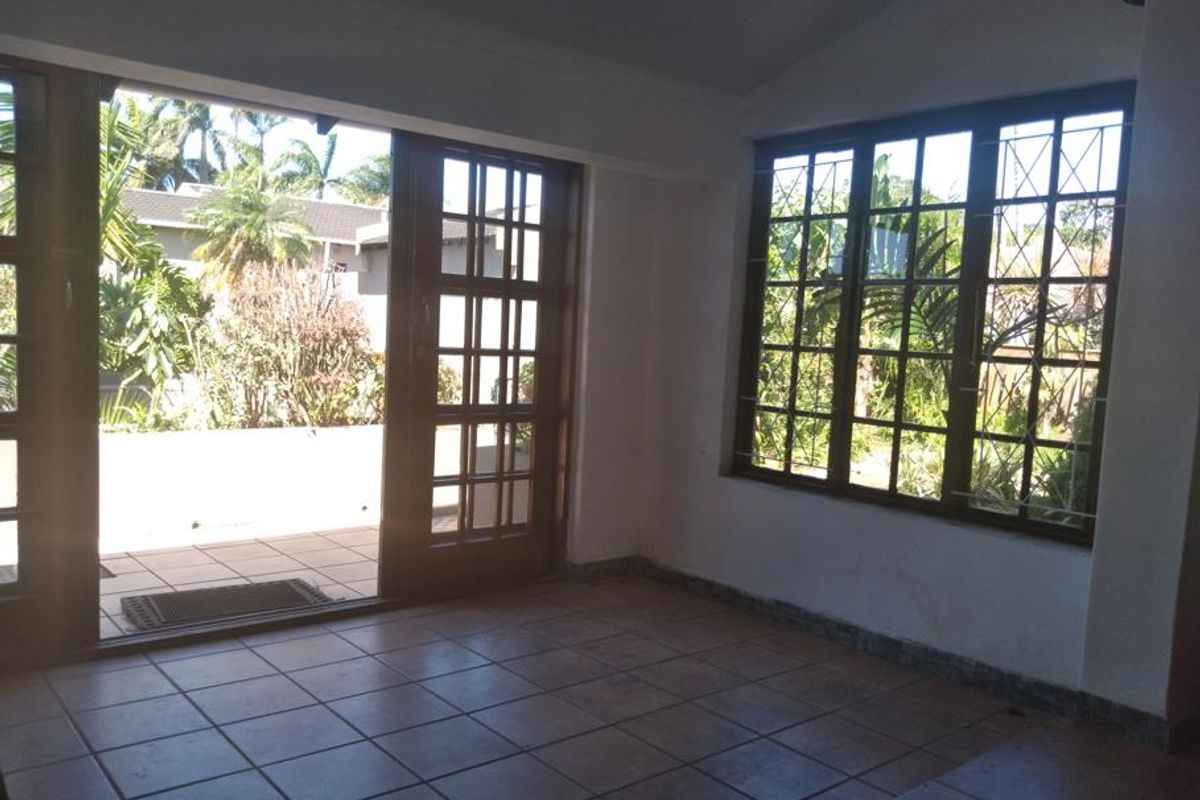 An empty living room featuring tiled flooring, timber-framed windows with security bars, and direct access to an outdoor patio area.