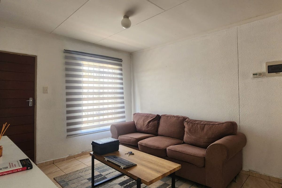 A compact living room featuring tiled flooring, neutral walls, and a window fitted with modern roller blinds.