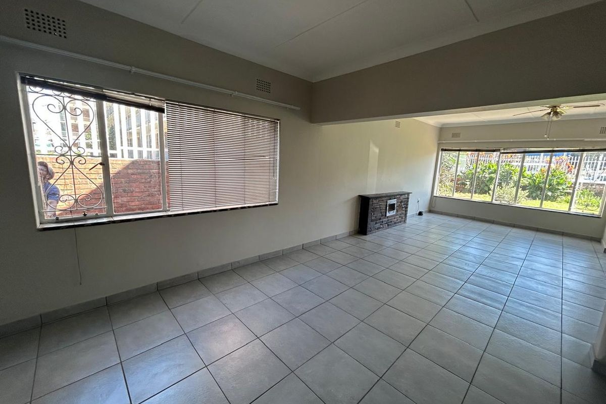 Spacious, light-filled living area featuring neutral tiled flooring, a brick fireplace, and large windows with security bars and blinds.