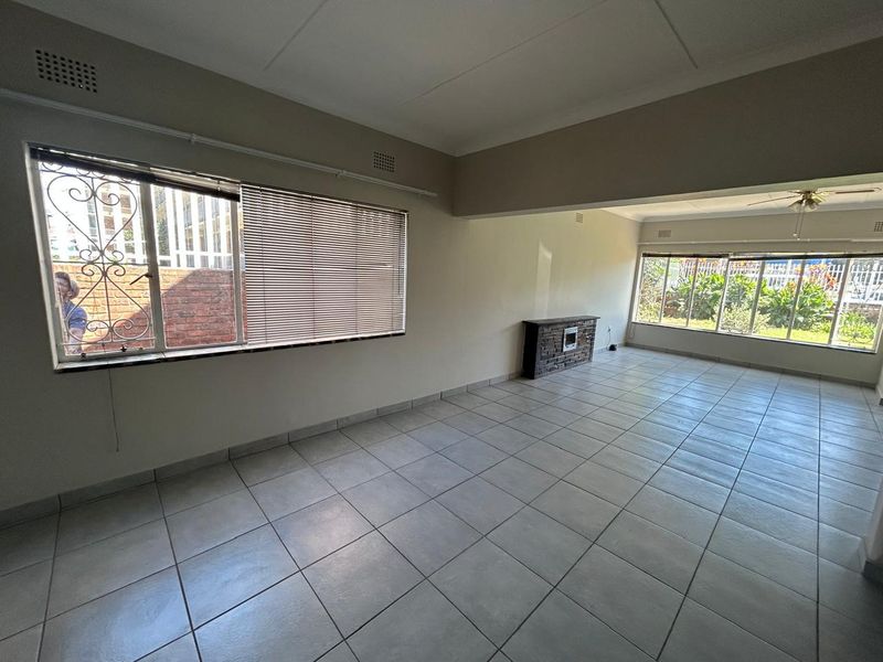 Spacious, light-filled living area featuring neutral tiled flooring, a brick fireplace, and large windows with security bars and blinds.
