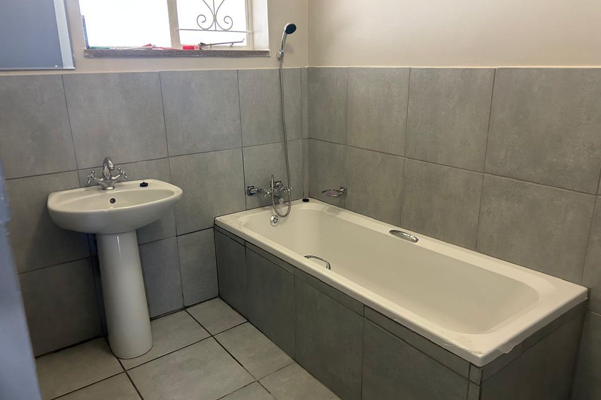 A clean, functional bathroom featuring a pedestal basin, a built-in bathtub with a handheld shower mixer, and neutral grey wall and floor tiling.