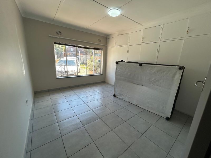 A bright, empty bedroom featuring neutral tiled flooring, built-in storage cupboards, and a window with security bars.