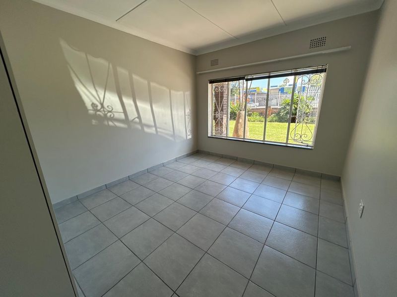 A bright, empty bedroom featuring neutral walls, tiled flooring, and security bars on the window overlooking the garden.